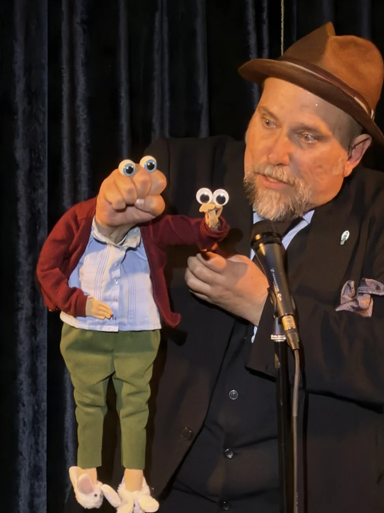 Ventriloquist Karl Herlinger in a black suit and brown fedora performs with his figure onstage in front of a curtain at the Peacock theater