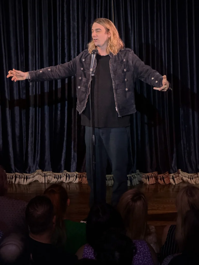 Marcus Monroe, with blond hair to his shoulders, a denim military-style jacket, black tee shirt and trousers, performs standup comedy in front of a microphone onstage in front of an audience at the Peacock theater