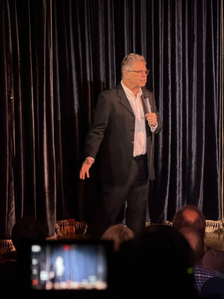Dana Gould, with grey hair and black glasses in a black suit, holds a microphone performing comedy onstage in front of an audience at the Peacock theater. The screen of a filming device can be seen, out of focus, in the audience.